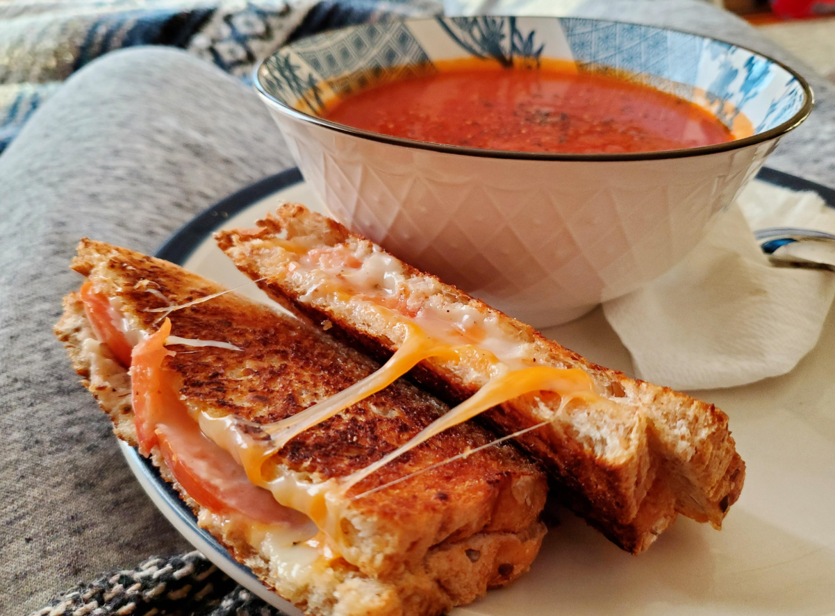 A bowl of tomato soup sits next to two halves of a grilled cheese sandwich with melted cheese on a plate, placed on a cozy blanket—perfect for enjoying comforting meals.