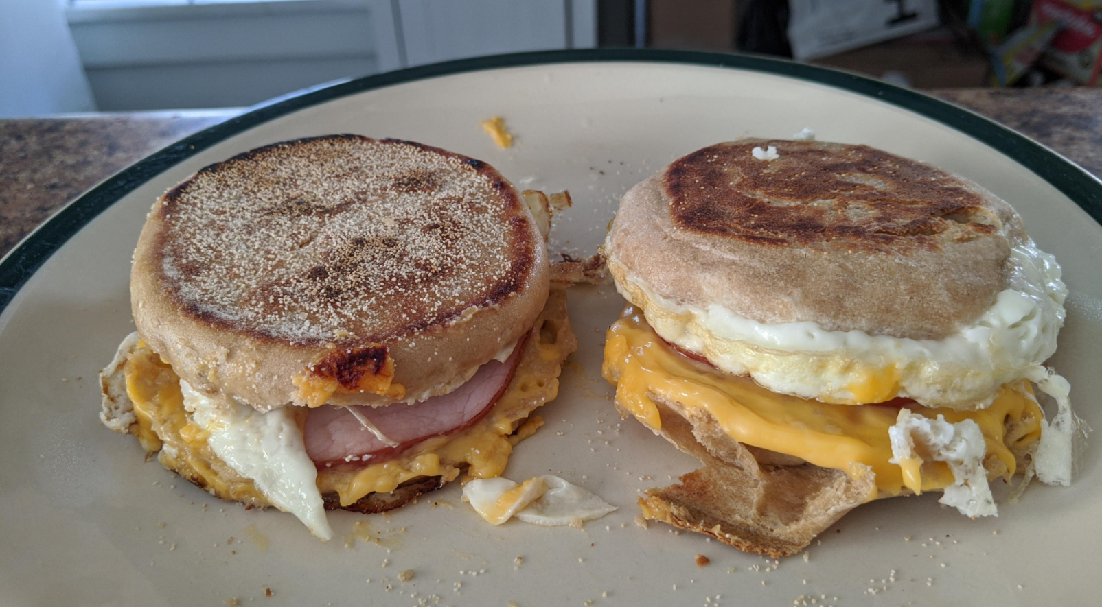 Two breakfast sandwiches on a plate, each made with an English muffin, egg, and melted cheese. One sandwich also has ham. The eggs and cheese slightly spill out from the sides, making these hearty meals perfect for starting your day.
