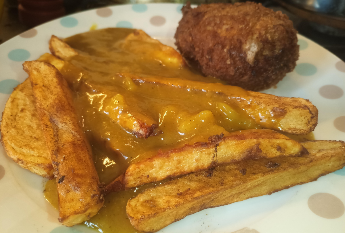 Thick-cut fries topped with curry sauce and a breaded, fried item—possibly a Scotch egg—served as one of the featured meals on a white plate with colored dots.