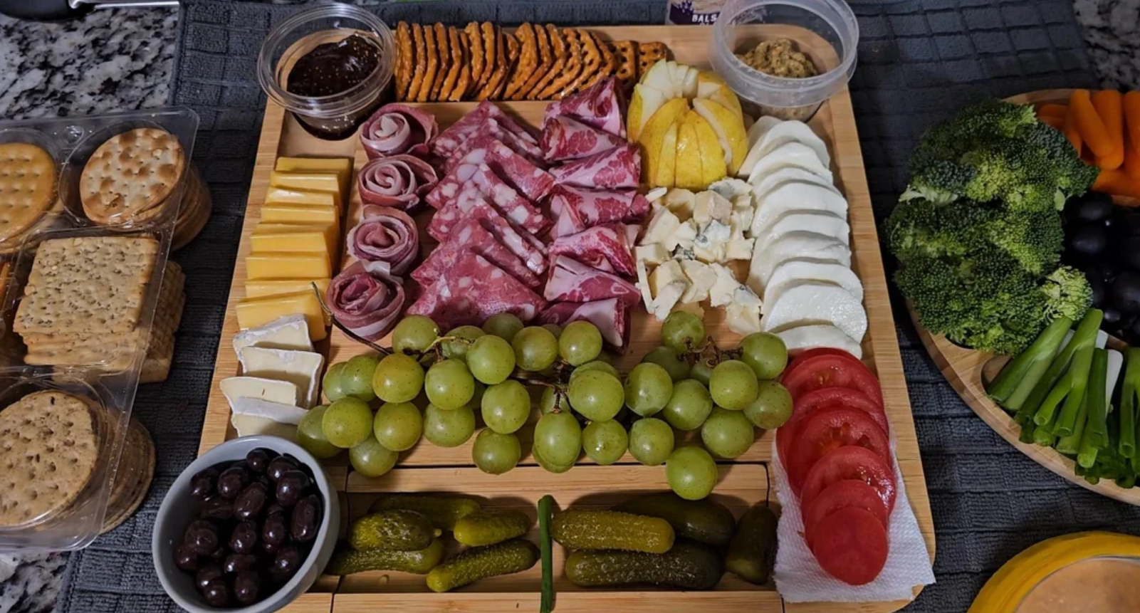 A charcuterie board with assorted cheeses, meats, grapes, crackers, olives, pickles, tomato slices, fig jam, and mustard is neatly arranged on a wooden board—perfect for sharing as part of casual meals or elegant gatherings.