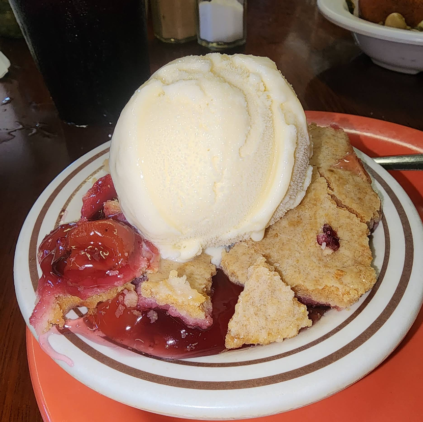 A plate of fruit cobbler topped with a large scoop of vanilla ice cream, served on a white plate with brown stripes, sits on an orange tray—just one sweet option you might find at a cheap all you can eat breakfast. A glass of dark beverage is in the background.