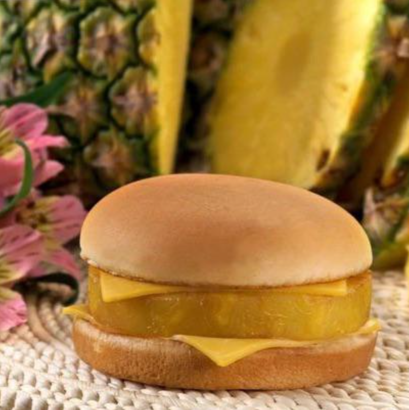 A sandwich bun filled with a slice of cheese and a thick pineapple ring sits on a table next to pink flowers and fresh pineapples in the background.