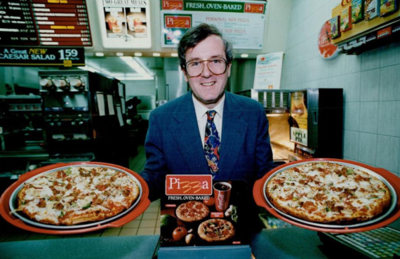 A man in a suit and tie stands behind a counter displaying two large pizzas in a restaurant, with menus and salad options visible in the background.