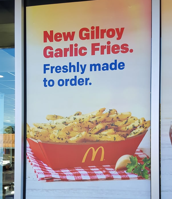 A McDonald’s sign advertises New Gilroy Garlic Fries with text and an image of seasoned fries in a red box, garnished with herbs and garlic cloves.
