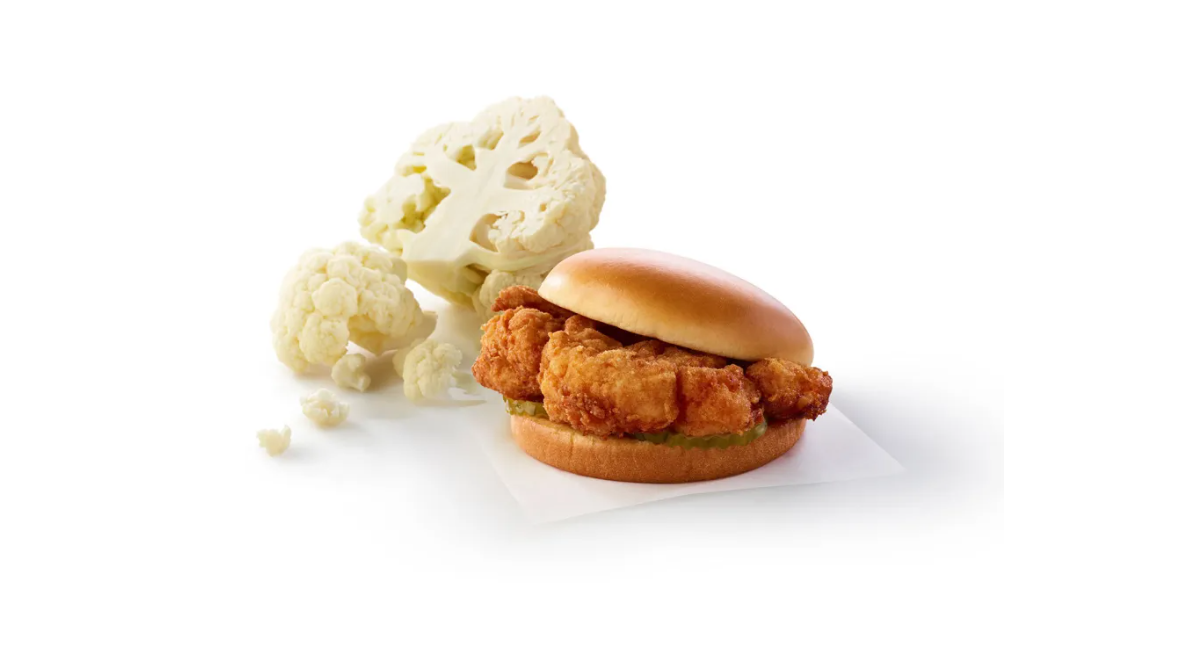 A fried cauliflower sandwich on a bun sits on white paper, with fresh cauliflower florets in the background, all set against a white surface.
