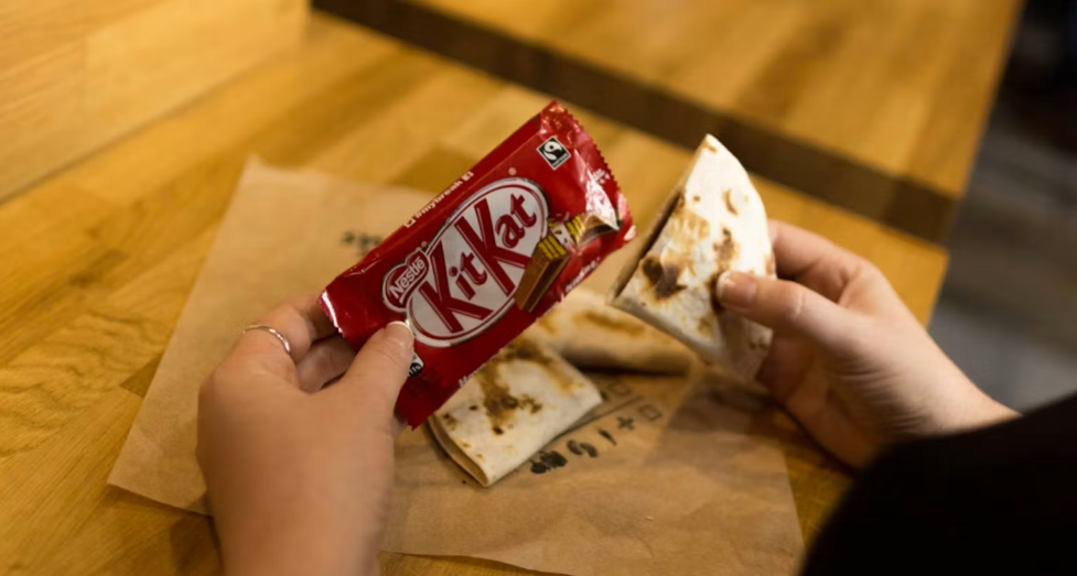 A person holds a KitKat bar and a quesadilla, with more quesadilla pieces on brown paper atop a wooden surface.