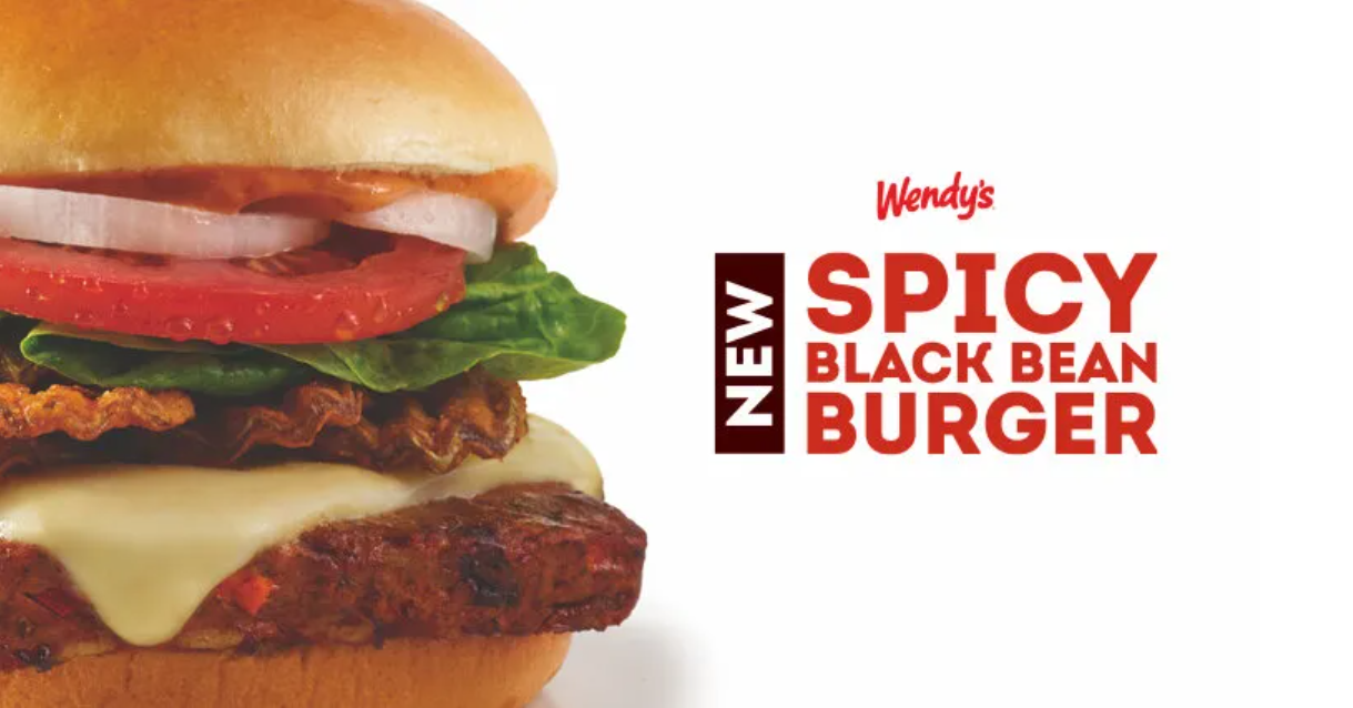 A Wendy’s spicy black bean burger with cheese, lettuce, tomato, onion, and sauce on a bun, next to text reading “NEW SPICY BLACK BEAN BURGER.”
