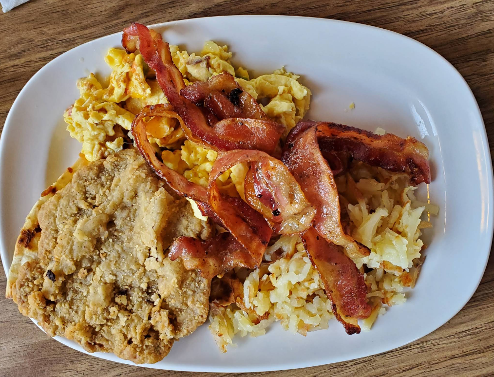 A white plate with scrambled eggs, crispy bacon strips, hash browns, and a breaded fried steak patty—all part of a cheap all you can eat breakfast on a wooden table.