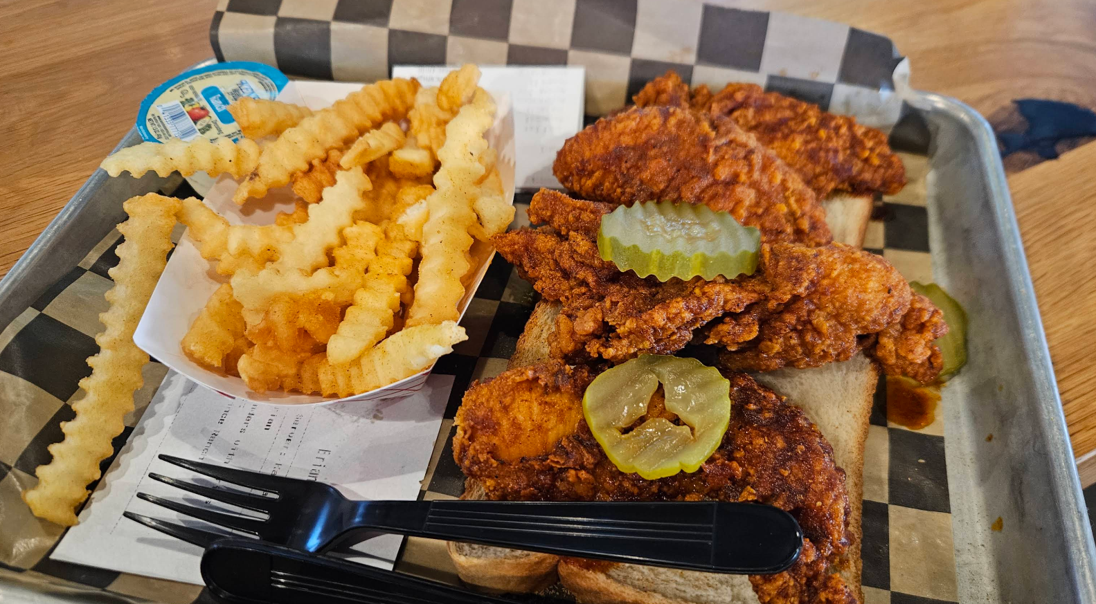 A tray with crinkle-cut fries in a paper cup, spicy fried chicken tenders on bread, pickle slices on top, a plastic fork, and a container of dipping sauce.