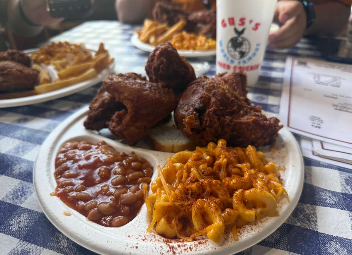 A plate of fried chicken, baked beans, and macaroni and cheese sits on a blue checkered tablecloth. In the background are fries, more food, and a Gus’s World Famous Fried Chicken cup.