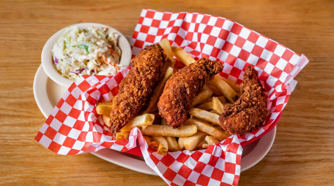 Three crispy fried chicken tenders and golden French fries are served in a basket lined with red and white checkered paper, with a side of creamy coleslaw in a small bowl on a wooden table.