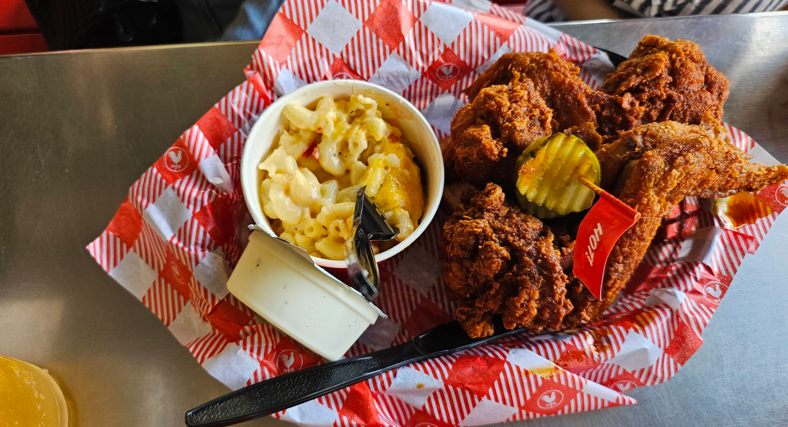 Basket with fried chicken topped with pickles, a small cup of macaroni and cheese, a white sauce container, black plastic fork and knife, all on red-and-white checkered paper on a metal table.