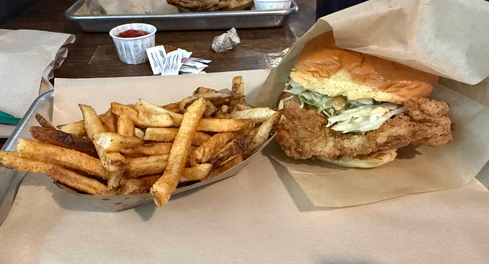 A fried chicken sandwich with lettuce on a bun sits next to a serving of golden French fries, both arranged on brown parchment paper.