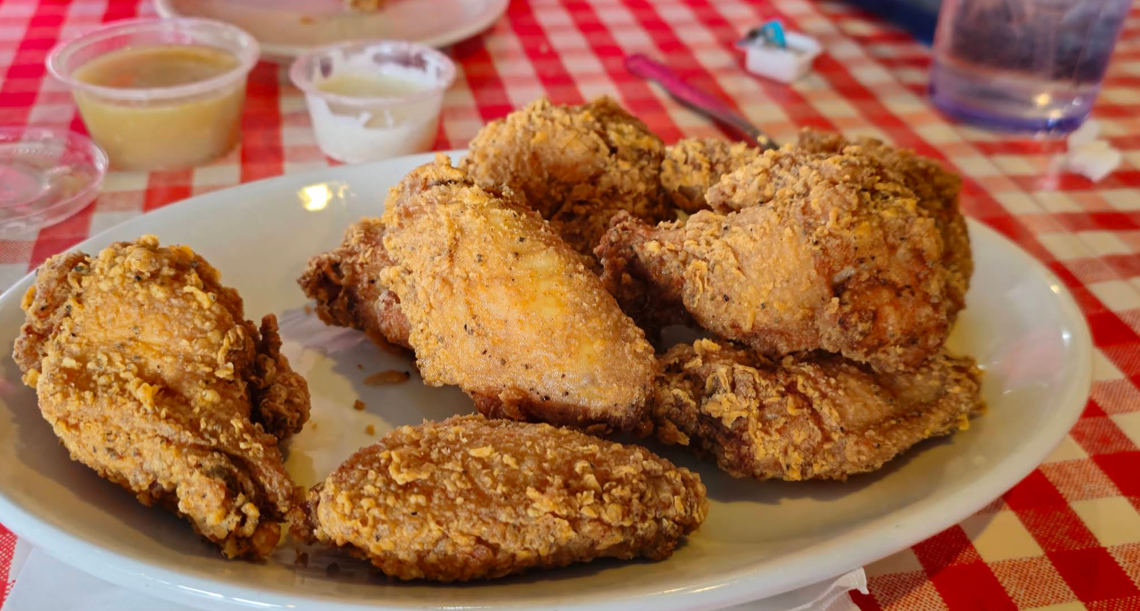 A white plate filled with pieces of crispy fried chicken sits on a red and white checkered tablecloth, with sauce containers and a drinking glass in the background.