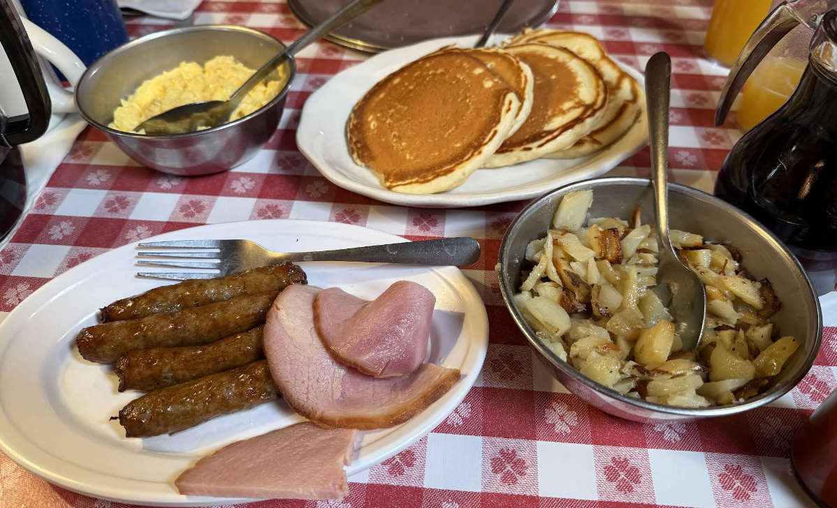A cheap all you can eat breakfast awaits on a red and white checkered tablecloth, featuring sausage links, ham slices, fried potatoes, scrambled eggs, pancakes, and a pitcher of syrup.