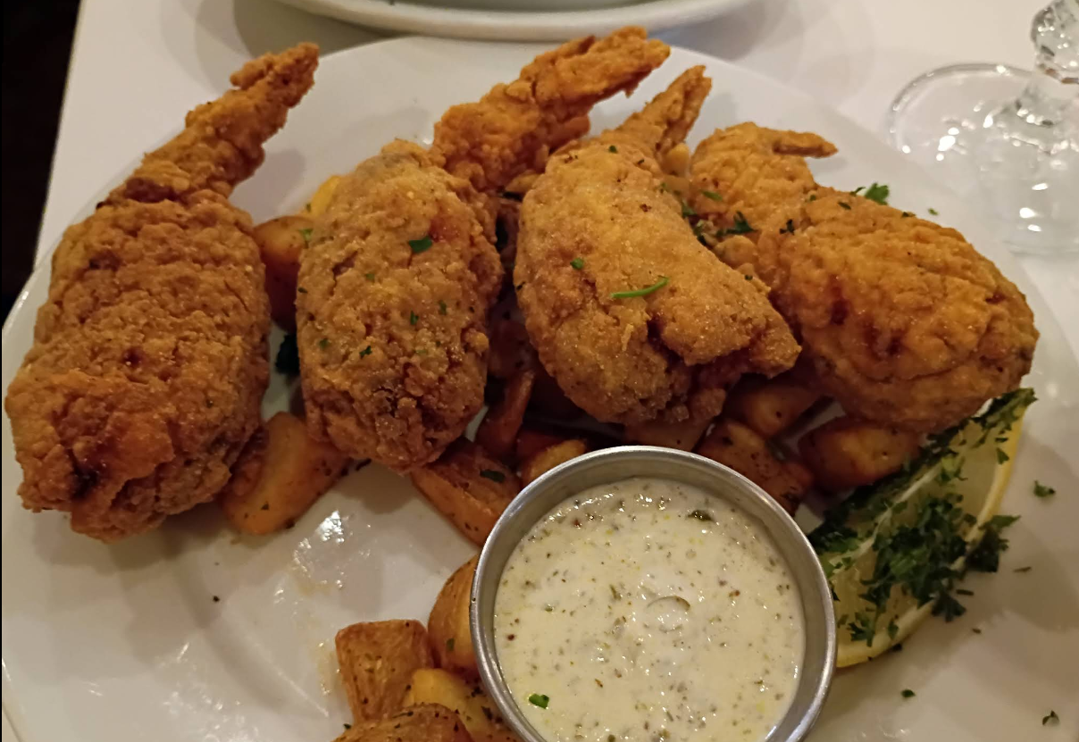 Four pieces of golden fried frog legs served on a bed of roasted potatoes, garnished with parsley, lemon wedge, and accompanied by a metal cup of creamy dipping sauce.