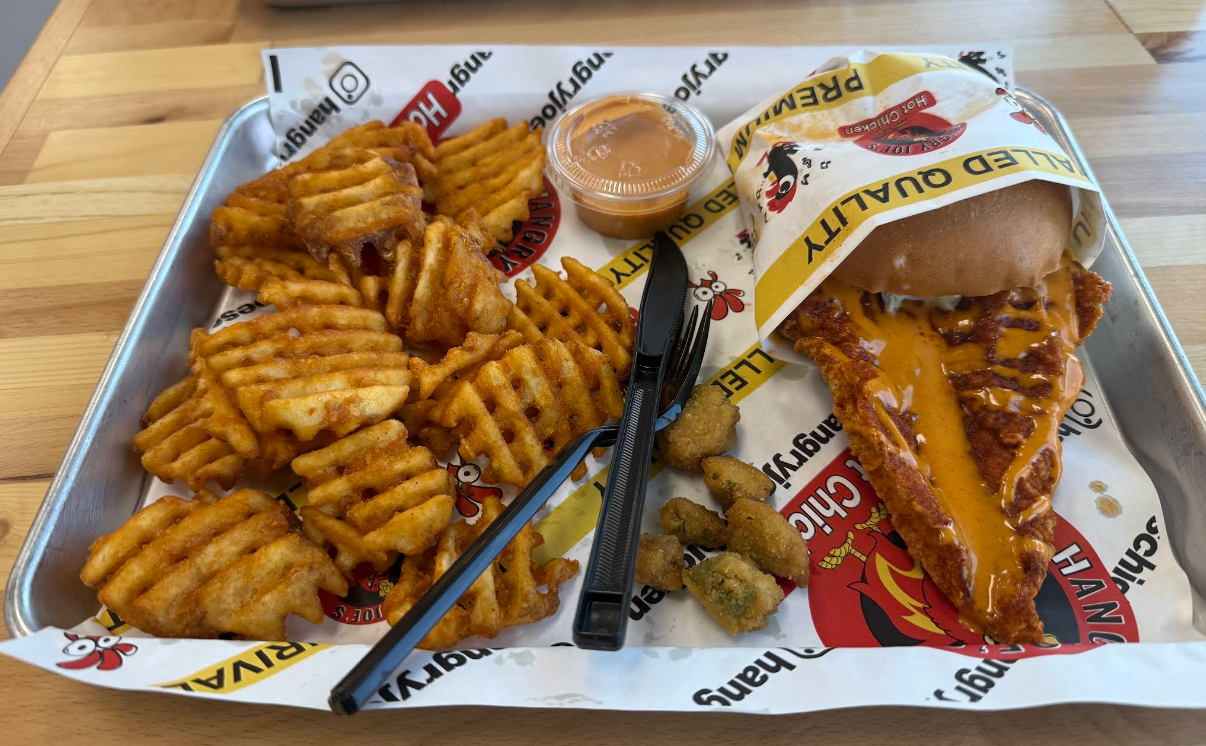 A tray with seasoned waffle fries, fried pickles, a spicy chicken sandwich with orange sauce, a cup of dipping sauce, and a black plastic fork and knife on branded paper lining—just the kind of mouthwatering meal you’d expect from one of the fastest-growing fried chicken chains.