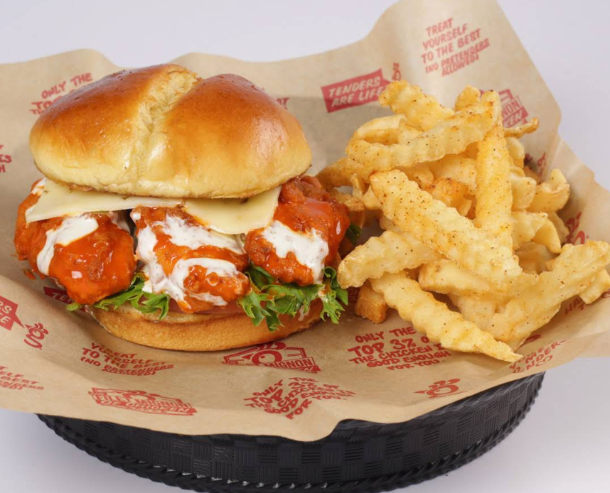A crispy fried chicken sandwich with lettuce, cheese, buffalo sauce, and ranch on a bun, served alongside crinkle-cut seasoned fries in a black basket lined with branded paper—a mouthwatering offering from one of the fastest-growing fried chicken chains.
