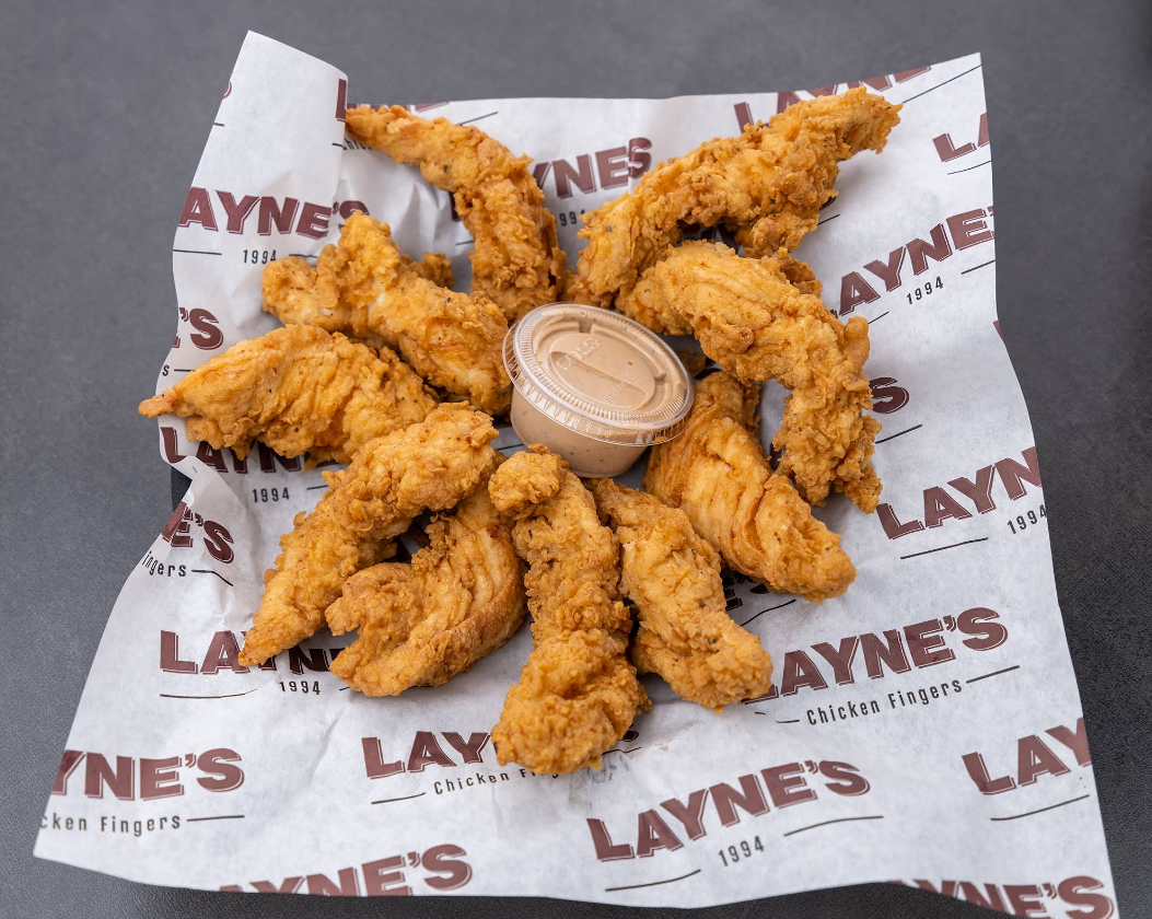 Eight crispy fried chicken tenders arranged in a circle around a container of dipping sauce, served on branded Layne's Chicken Fingers paper atop a gray surface—a mouthwatering offering from one of the fastest-growing fried chicken chains.