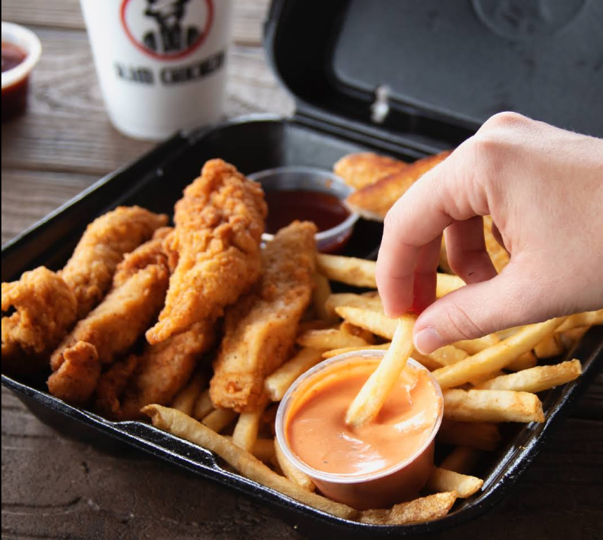 A hand dips a French fry into orange sauce in a black takeout box with chicken tenders, fries, and dipping sauces from one of the fastest-growing fried chicken chains. A drink cup sits in the background on a wooden table.