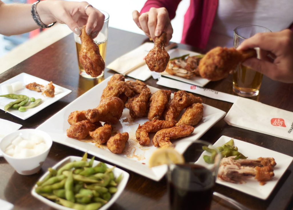 Three people grab pieces of fried chicken from plates on a table, surrounded by side dishes like edamame and drinks—a scene reminiscent of the lively atmosphere found at some of the fastest-growing fried chicken chains.