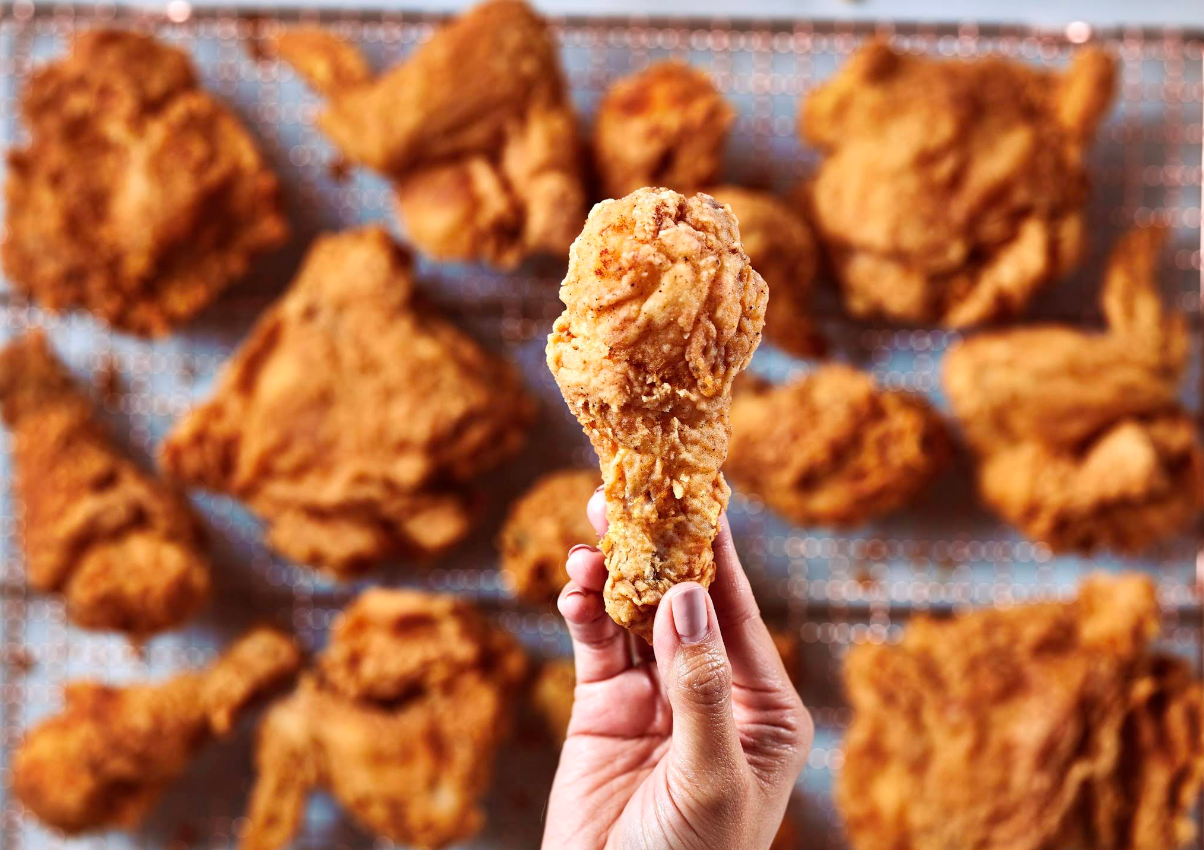 A hand holds a piece of crispy fried chicken drumstick above a wire rack filled with assorted fried chicken pieces in the background, capturing the irresistible appeal of one of the fastest-growing fried chicken chains.