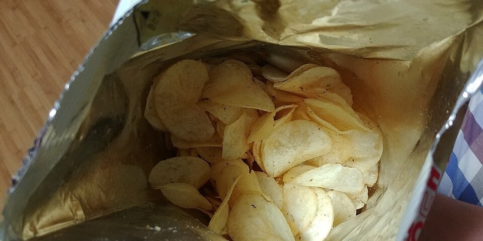 Open bag of potato chips viewed from above, showing several yellow, thin chips inside a shiny, foil-lined bag. Part of a plaid surface and wooden floor are visible at the edges.
