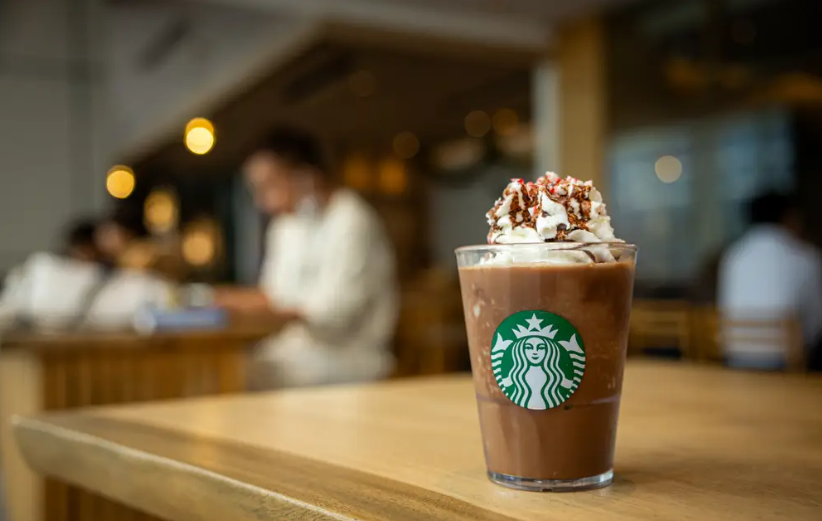 A Starbucks iced chocolate drink topped with whipped cream and chocolate drizzle sits on a wooden table inside one of the cozy coffee houses, with blurred people and warm lights in the background.