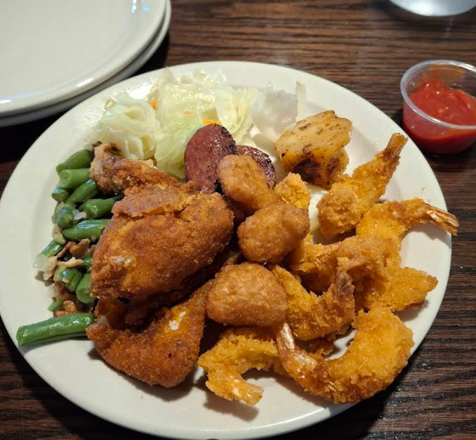 A plate of fried shrimp, fried chicken, hush puppies, green beans, sausage, cabbage, and potatoes—with a small cup of cocktail sauce—served on a wooden table at a cheap all you can eat breakfast spot.