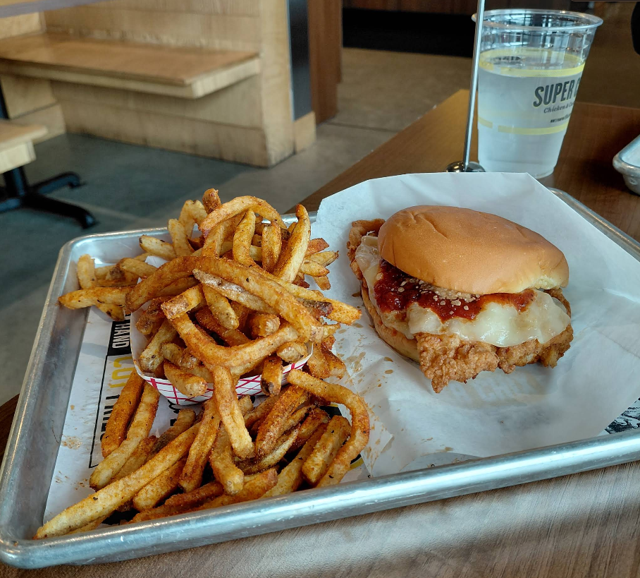 A tray with seasoned fries in a paper tray and a fried chicken sandwich with cheese and sauce, next to a plastic cup of water on a wooden table in one of the fastest-growing fried chicken chains' casual restaurant.