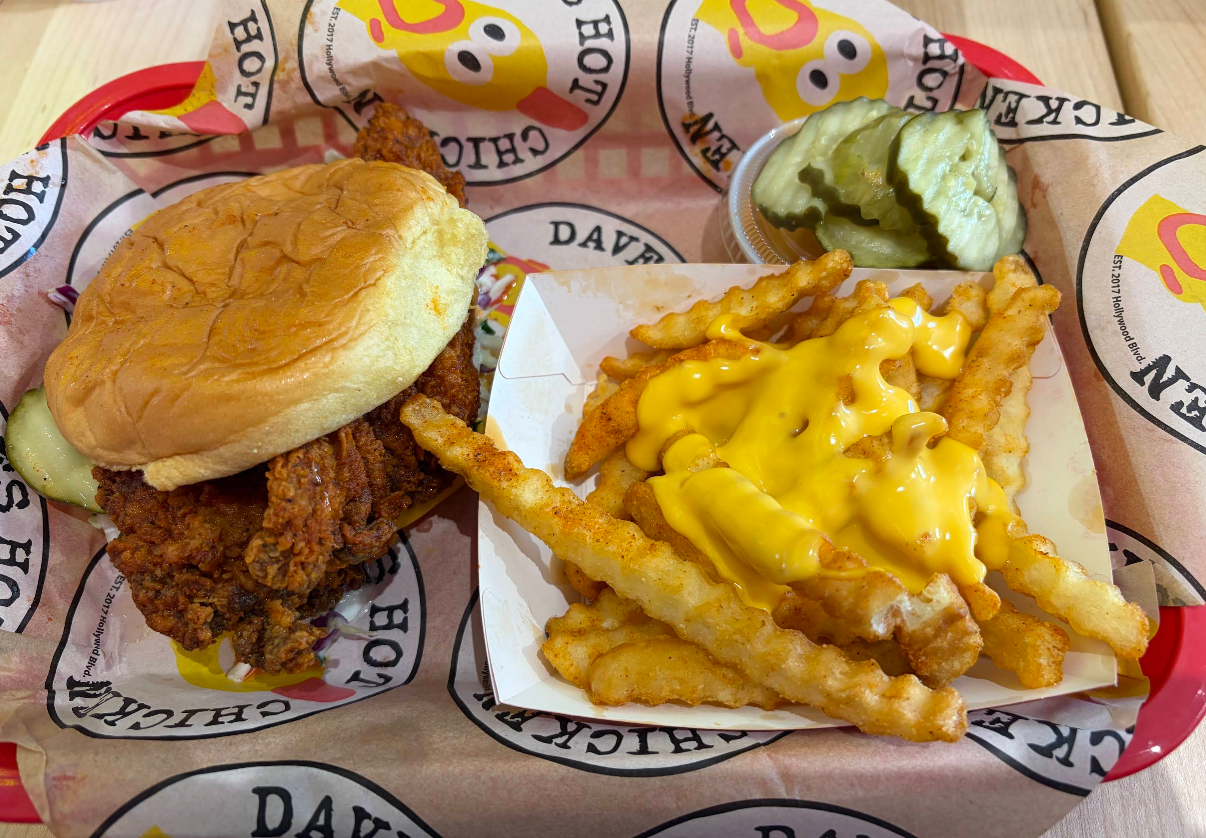A basket with a fried chicken sandwich, crinkle-cut cheese fries, and a small side of pickles, served on branded paper from one of the fastest-growing fried chicken chains.