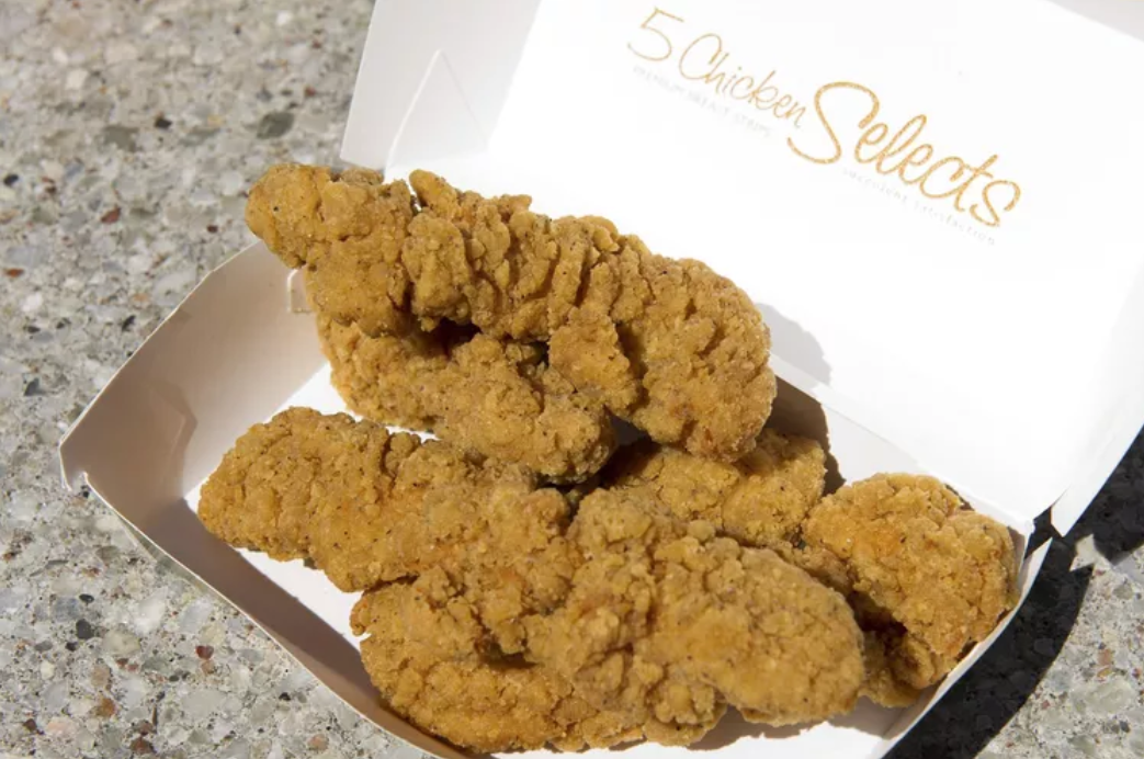 Four crispy, golden-brown chicken tenders in an open white box labeled "5 Chicken Selects" on a speckled countertop.