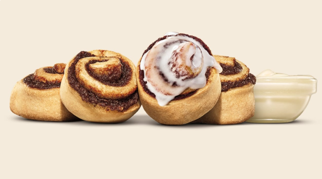 Four cinnamon rolls are displayed, one topped with white icing. A small clear bowl of creamy white icing sits to the right, all set against a plain light background.
