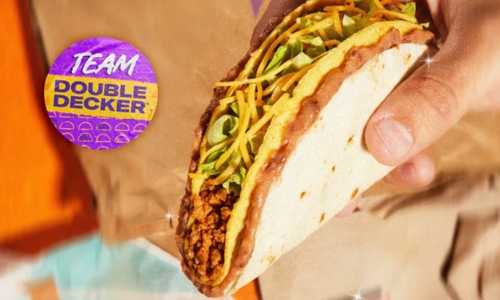 A hand holds a Double Decker taco filled with meat, lettuce, and cheese. A purple badge reads “TEAM DOUBLE DECKER” on the left side. The background is orange and brown.