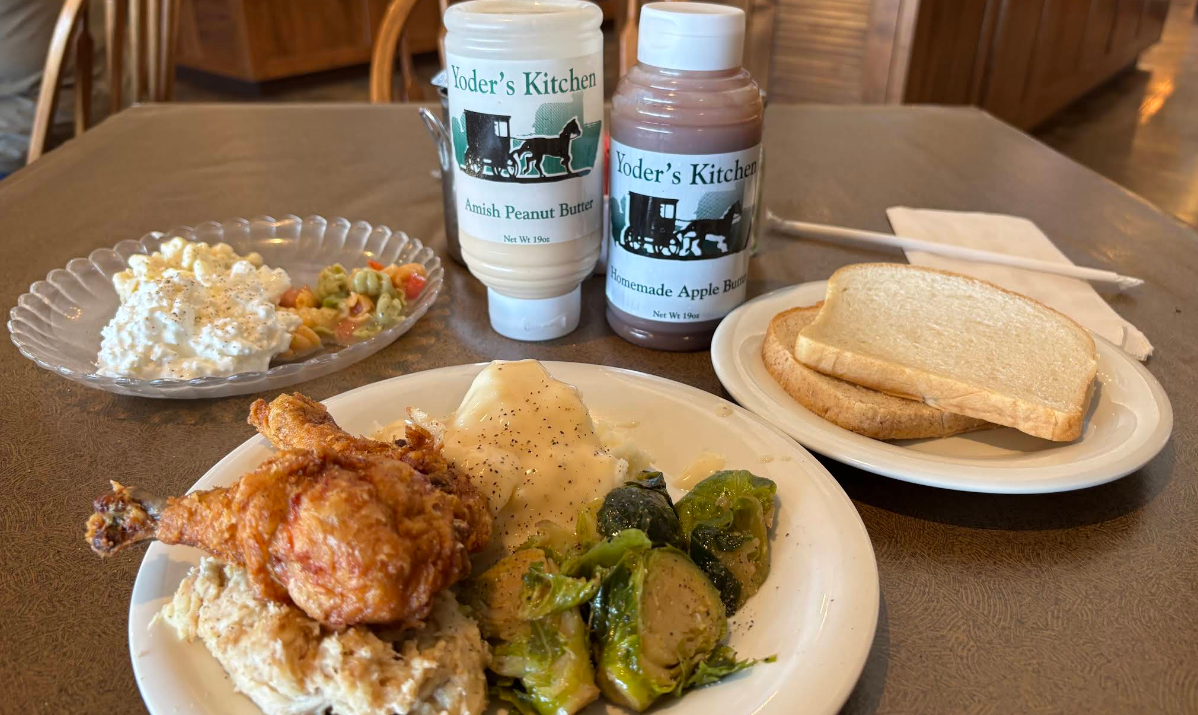 A table set for a cheap all you can eat breakfast features fried chicken, mashed potatoes with gravy, Brussels sprouts, noodles, a fresh vegetable salad, slices of white bread, and bottles of Amish peanut butter and homemade apple butter.