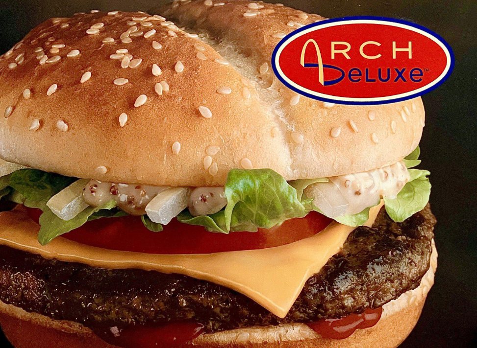 A close-up of a hamburger with lettuce, tomato, onions, cheese, and sauce on a sesame seed bun. The "Arch Deluxe" logo appears in the top right corner.