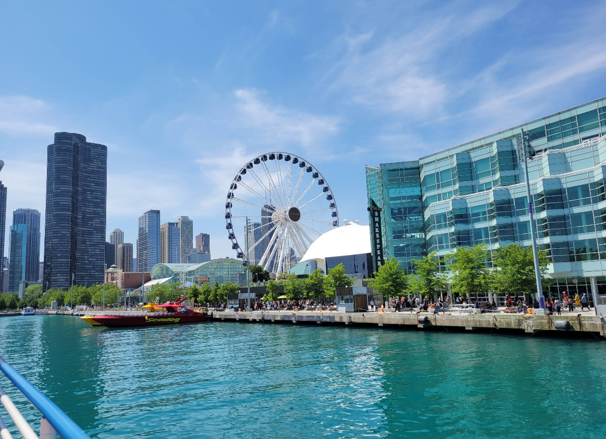 A large Ferris wheel stands near modern glass buildings by the waterfront, making it one of the city's iconic destinations, with a red and yellow speedboat in the blue water and a city skyline in the background under a partly cloudy sky.