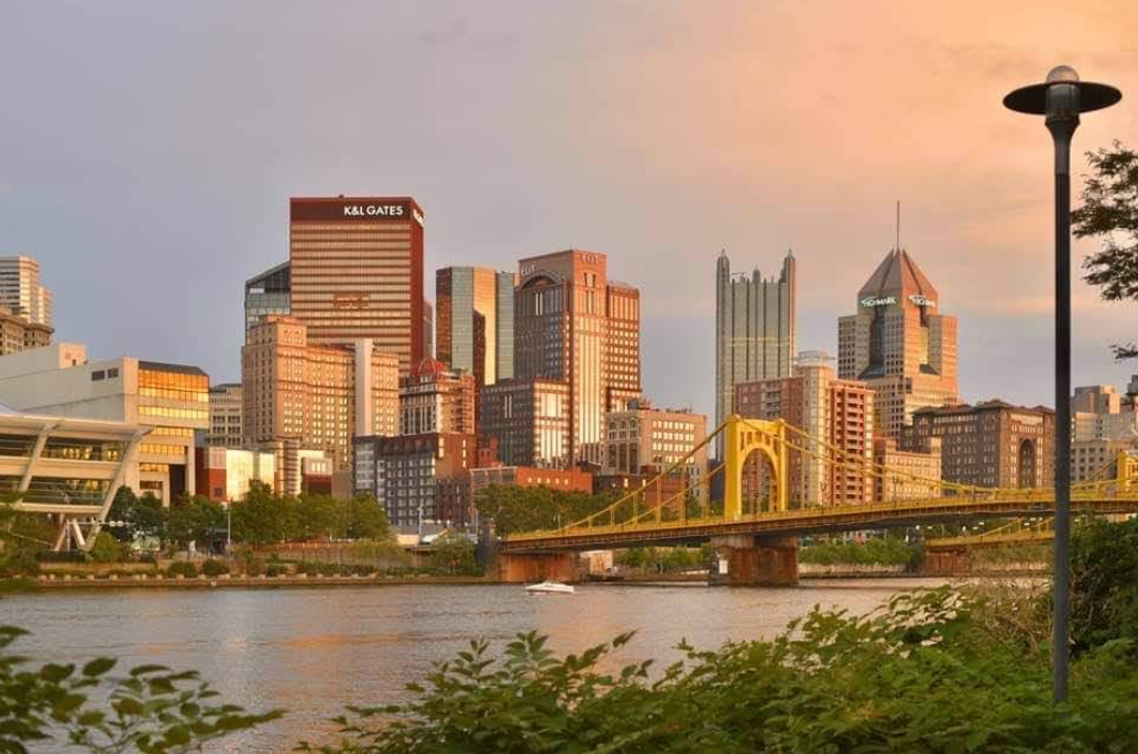 A stunning view of the Pittsburgh skyline at sunset showcases iconic skyscrapers and the yellow Roberto Clemente Bridge spanning the river in the foreground. As a streetlamp glows and lush greenery frames the scene, this picturesque cityscape highlights one of Pennsylvania’s must-see destinations.