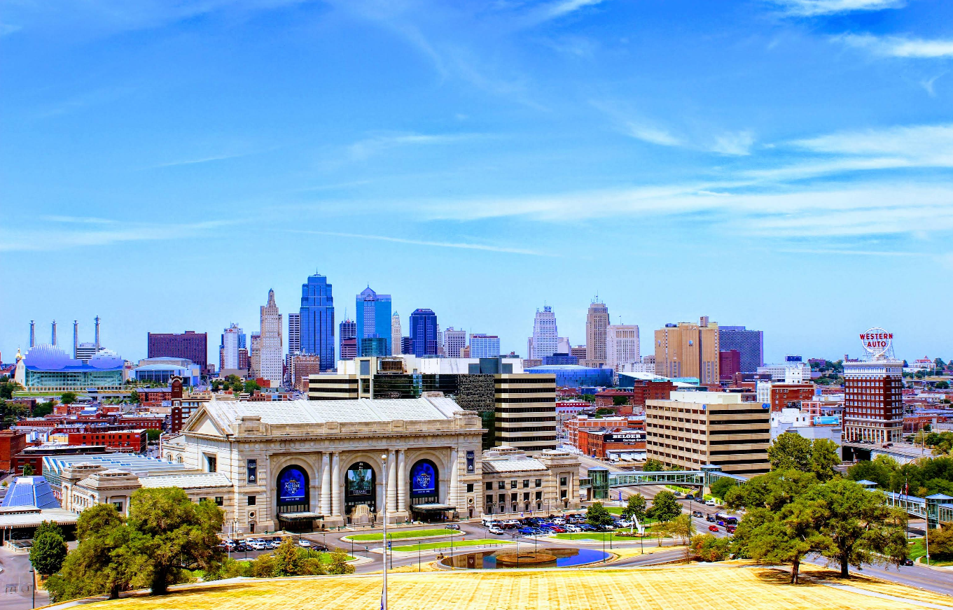 Kansas City skyline featuring Union Station in the foreground, with modern office buildings and skyscrapers in the background beneath a bright blue sky with scattered clouds. Trees and green spaces are also visible, making this cityscape one of the must-see destinations for travelers seeking a blend of history, modernity, and vibrant urban scenery.