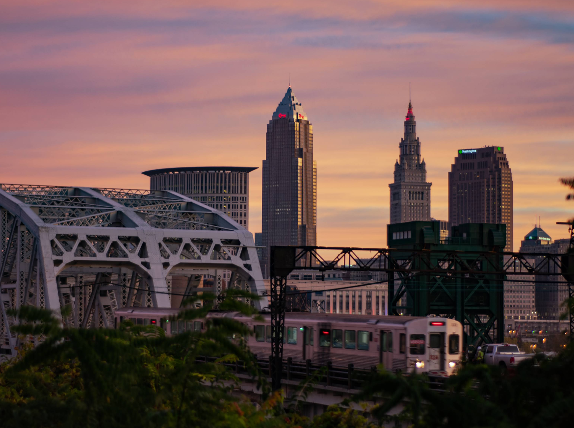 A city skyline at sunset with tall buildings, a metal bridge in the foreground, and a train passing through—reminding travelers of the endless destinations waiting just beyond the horizon. Soft pink and orange clouds color the sky above.