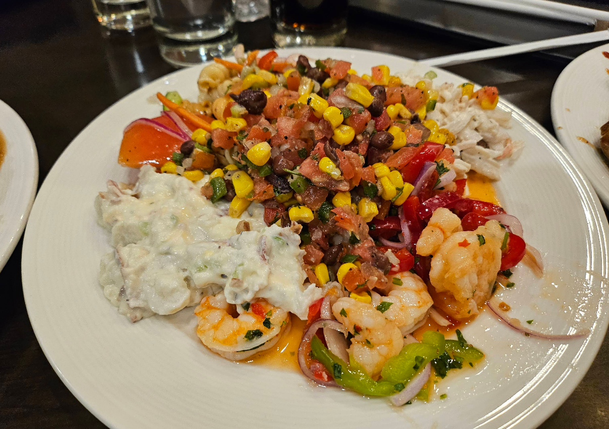 A plate of mixed seafood salad with shrimp, chopped vegetables, corn, black beans, guacamole, and pico de gallo, served cold on a white dish—perfect before a cheap all you can eat breakfast. Glasses of drinks are visible in the background.
