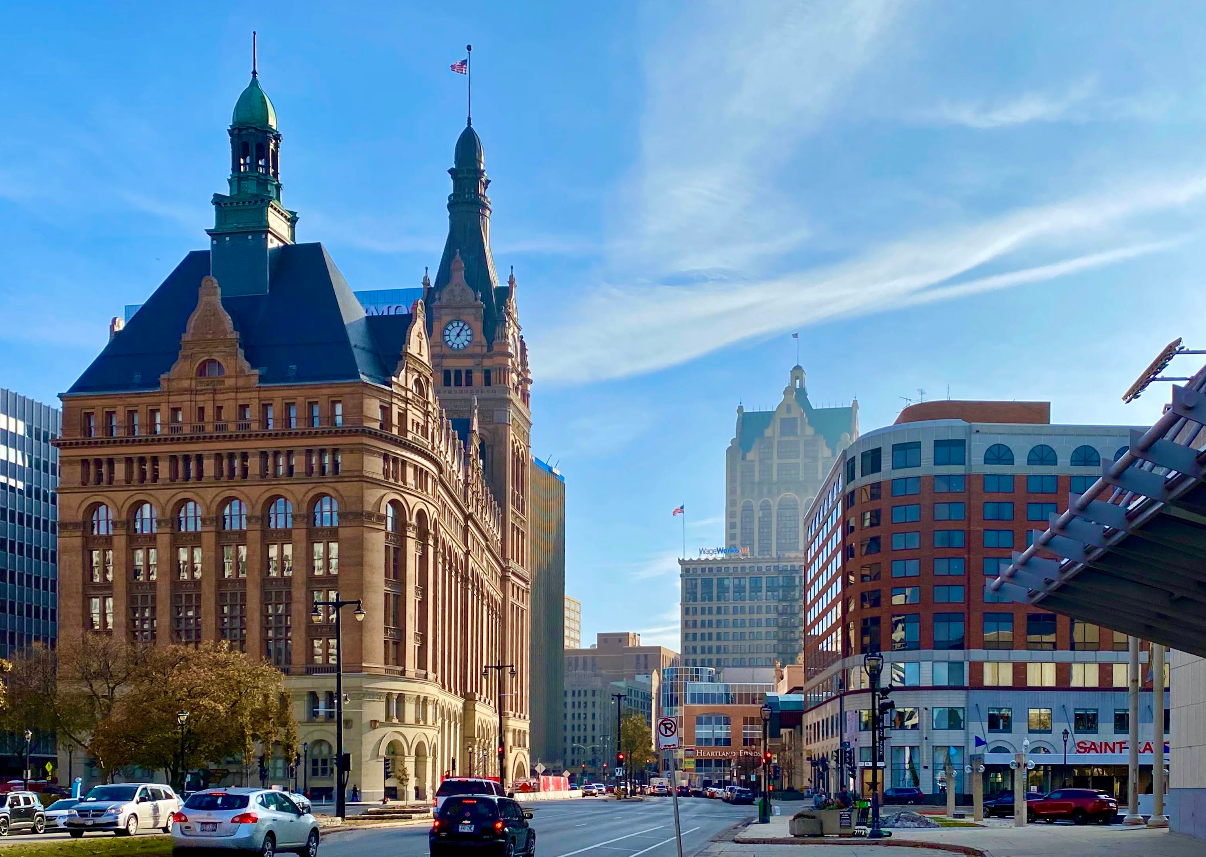 Street view of downtown Milwaukee with historic buildings, including the Milwaukee City Hall with its clock tower, cars, and a clear blue sky—highlighting one of the city's must-see destinations.