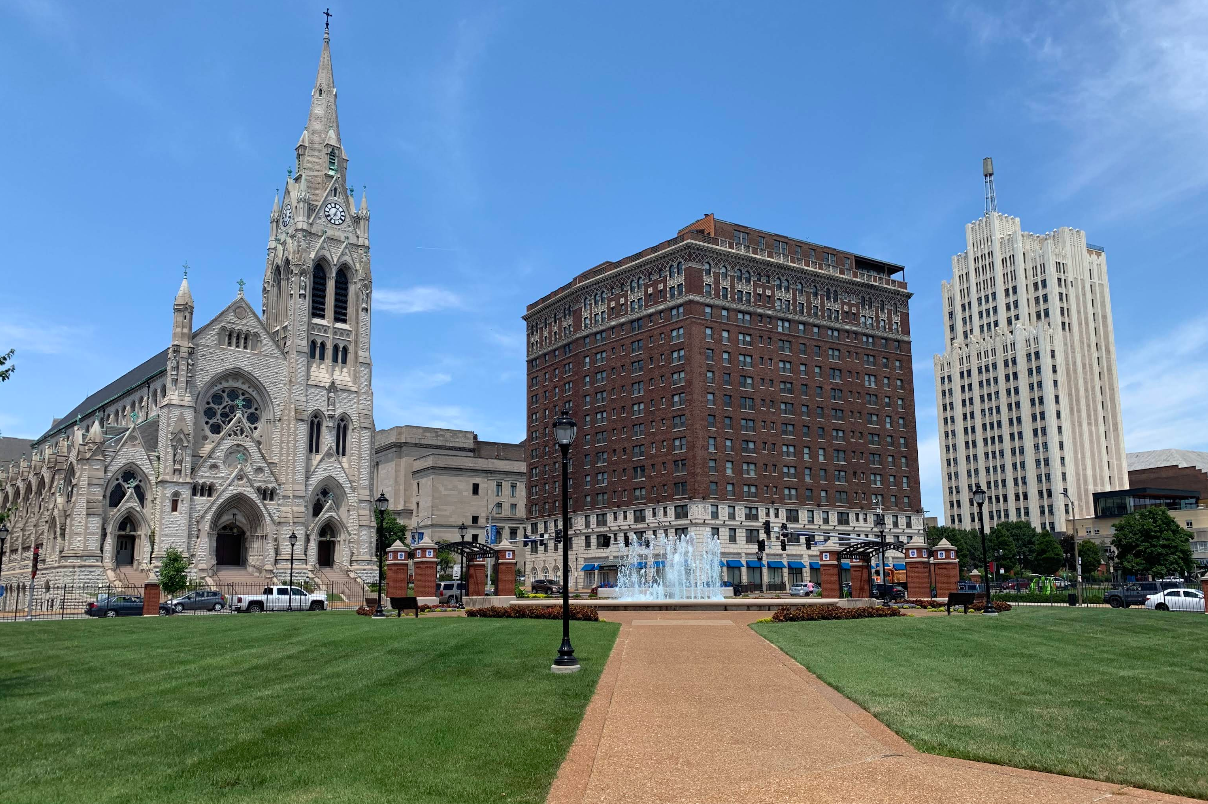 A large church, a brown brick building, and a white tower stand near a fountain in a grassy park under a blue sky, making this area one of the must-see destinations for visitors.