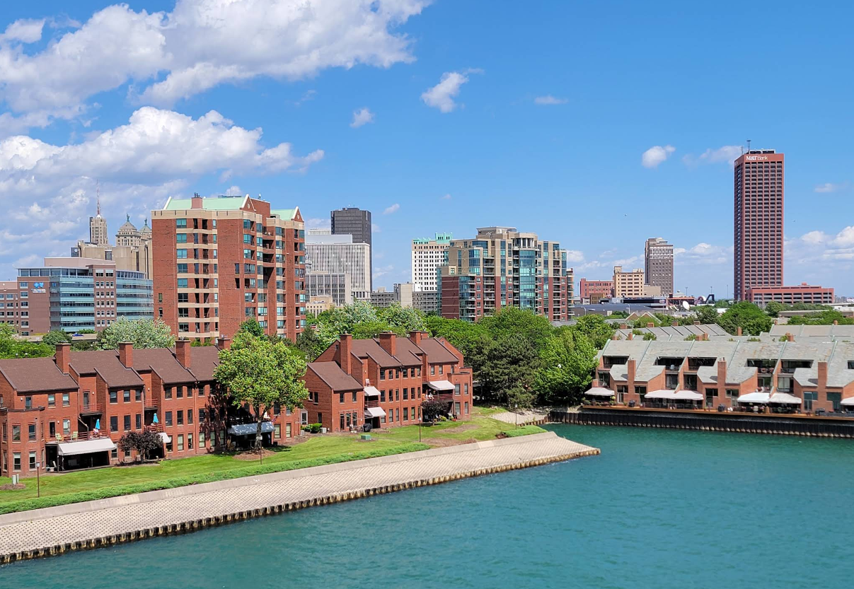 Brick buildings and townhouses line a riverbank in front of a city skyline with tall modern and historic buildings, making this area one of the most picturesque destinations to explore under a bright blue sky with scattered clouds.