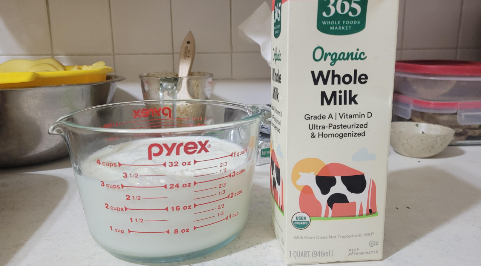A measuring cup containing milk sits next to a carton of Whole Foods Market Organic Whole Milk on a kitchen counter with bowls and utensils in the background.