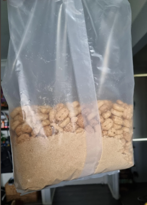 A close-up of a plastic cereal bag shows a large amount of crushed powder at the bottom, with some whole cereal pieces above it. The background includes shelves and a door inside a home.