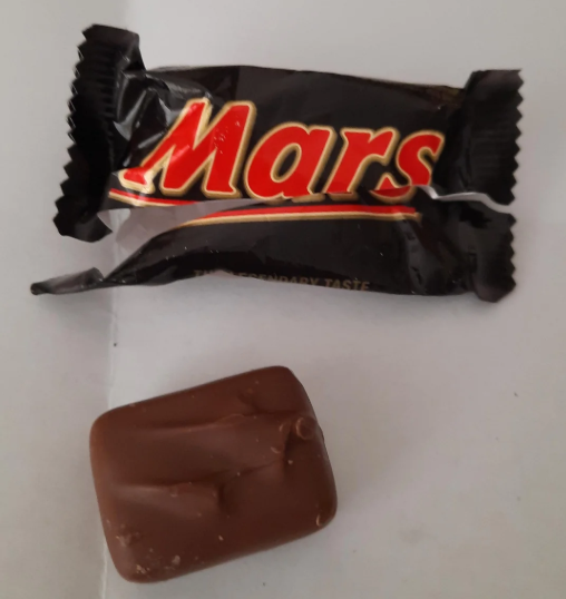 A Mars chocolate bar in black packaging is placed above an unwrapped piece of the bar, showing its smooth chocolate coating, all on a white surface.