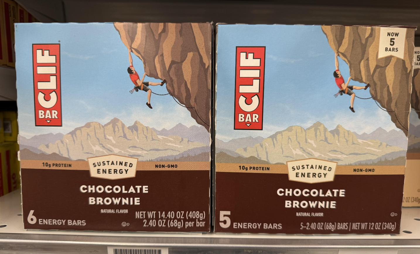 Two boxes of Clif Bar Chocolate Brownie energy bars on a store shelf, each featuring a climber scaling a cliff with mountains in the background. The boxes display protein content and number of bars inside.