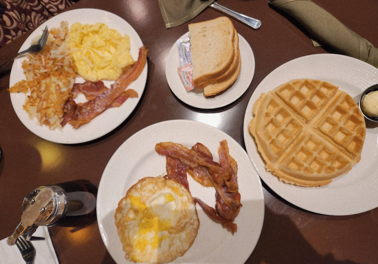 A cheap all you can eat breakfast spread with scrambled eggs, hash browns, bacon, toast, waffles with butter, an egg, and a small bottle of syrup on a brown table.