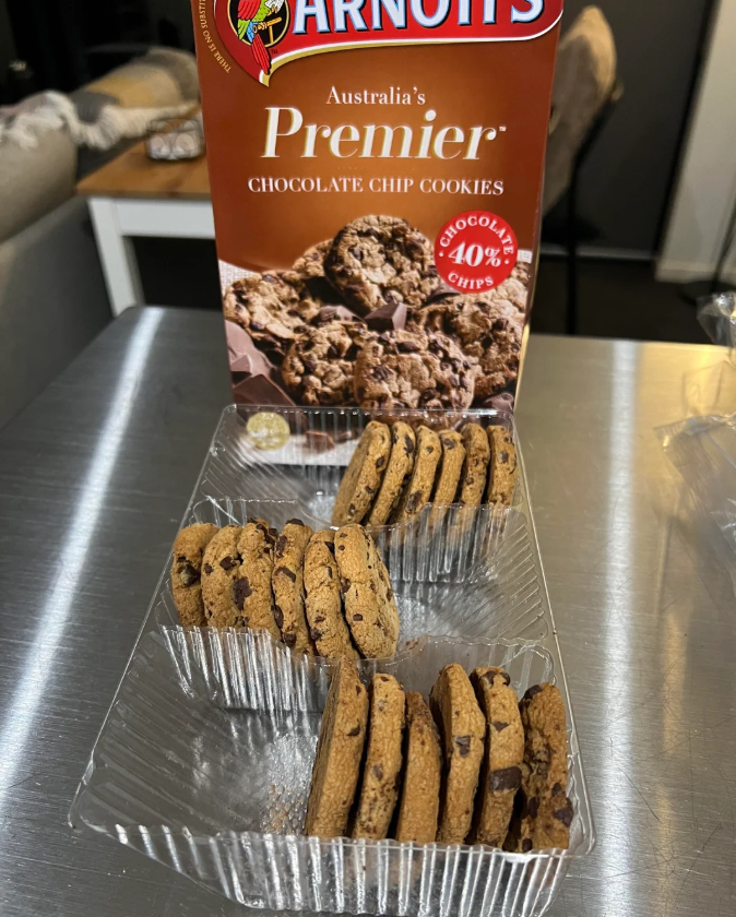 An open plastic tray of Arnott's Premier Chocolate Chip Cookies sits on a metal surface, with several cookies standing upright. The packaging behind the tray displays the brand and an image of cookies.
